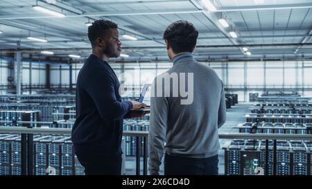Two Multiethnic Male Data Center System Administrators and IT Specialists Talk, Use Laptop. Information Technology Engineers work on Cyber Security Network Protection in Cloud Computing Server Farm. Stock Photo