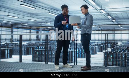 Two Multiethnic Male Data Center System Administrators and IT Specialists Talk, Use Laptop. Information Technology Engineers work on Cyber Security Network Protection in Cloud Computing Server Farm. Stock Photo