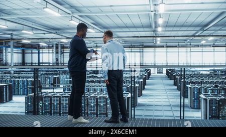 Male Data Center System Administrators and African American IT Specialists Talk, Use Laptop. Information Technology Engineers work on Cyber Security Network Protection in Cloud Computing Server Farm. Stock Photo