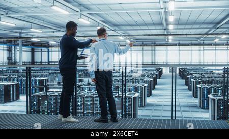 Male Data Center System Administrators and African American IT Specialists Talk, Use Laptop. Information Technology Engineers work on Cyber Security Network Protection in Cloud Computing Server Farm. Stock Photo