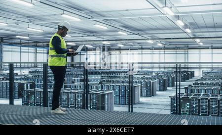 Data Center Engineer in High Visibility Vest Using Laptop Computer. Server Farm Cloud Computing Facility with Black Male System Administrator Working with Data Protection Network for Cyber Security. Stock Photo
