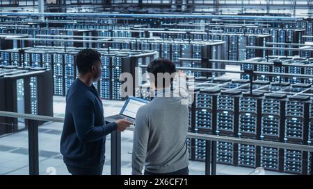 Two Multiethnic Male Data Center System Administrators and IT Specialists Talk, Use Laptop. Information Technology Engineers work on Cyber Security Network Protection in Cloud Computing Server Farm. Stock Photo