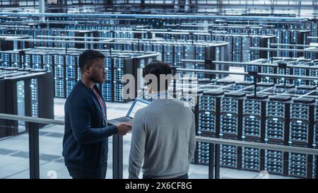 Two Multiethnic Male Data Center System Administrators and IT Specialists Talk, Use Laptop. Information Technology Engineers work on Cyber Security Network Protection in Cloud Computing Server Farm. Stock Photo