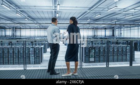 Data Center Female System Administrator and Male IT Specialist Talk, Use Laptop. Information Technology Engineers work on Cyber Security Network Protection in Cloud Computing Server Farm. Stock Photo