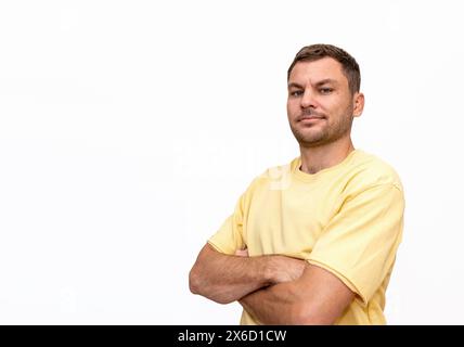 Isolated portrait of man wearing yellow t-shirt standing with crossed arms, smiling, and looking at camera. Isolated on white background. Stock Photo