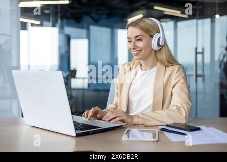 A smiling woman programmer wearing headphones while working on a laptop in a modern office environment, representing focus and productivity. Stock Photo