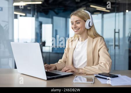 A smiling woman in a beige blazer works on a laptop in a glass-walled office, wearing headphones and enjoying her work. Stock Photo
