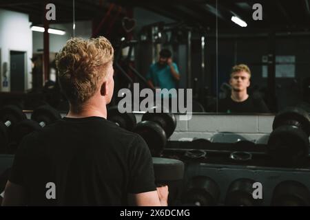 Rear view shot of a sportsman holding dumbbells, resting during his workout, sitting in front of gym mirror Stock Photo