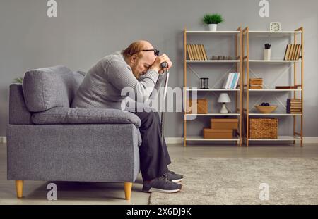 Red-haired, thoughtful, elderly man in gray warm sweater bowed his head, leaning on cane, sitting on gray sofa at home or in nursing home. Care for Stock Photo