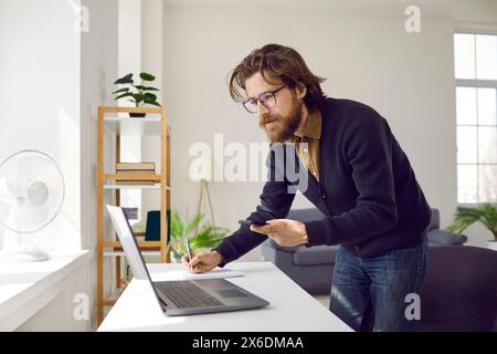 Hurrying businessman working from home writing making notes looking at laptop and smartphone. Stock Photo