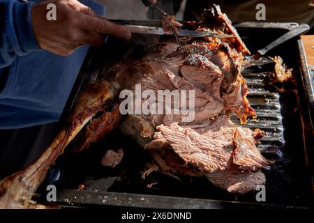 man's hands cutting a leg of lamb with a knife on a grill in Argentina Stock Photo