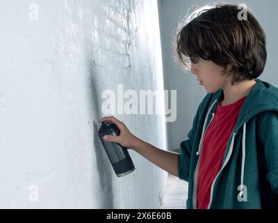A boy is spraying paint on a wall. The boy is wearing a red shirt and a green hoodie Stock Photo