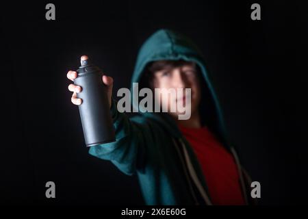 A young man holds a can of spray paint in his hand pointing to start his graffiti. He is wearing a green hooded sweatshirt and a red t-shirt. Concept Stock Photo