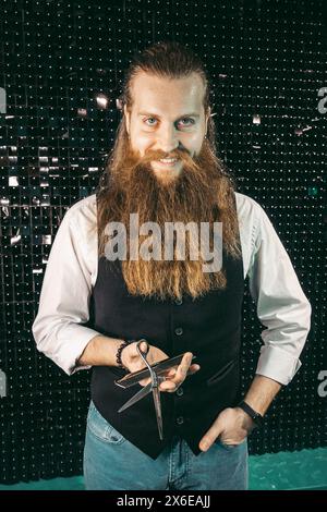Bearded barber portrait with scissors .Man young barber hairdresser professional with long beard and hair in vest with scissors in hands portrait, cut Stock Photo