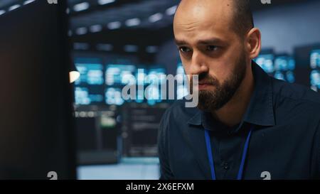 Overworked engineer feeling stressed while coding in data center facility, trying to ensure optimal performance of supercomputers. Tired man feeling fatigued while using PC to monitor server cabinets Stock Photo