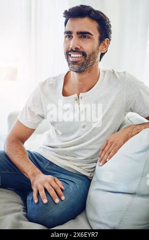 Indian man, thinking and happy or relax on sofa with daydreaming, comfortable and weekend break. Person, smile and thoughtful on couch in house with Stock Photo