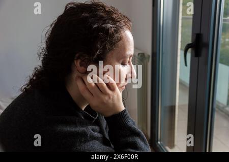 Close-up of sad woman with her head down looking through the window with her eyes closed Stock Photo