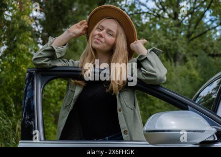 Blonde woman in hat staying next to car door. Young tourist explore local travel making candid real moments. True emotions expressions of getting away and refresh relax on open clean air Stock Photo