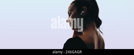 Woman with a ponytail hairstyle stands in a studio setting, using wireless earbuds to listen to music. She looks away, appearing relaxed and peaceful. Stock Photo