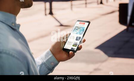 Close Up of a Young Man Using Smartphone for Scrolling News on a Street During a Sunny Day. He is Reading Latest News About Science. Display in Vertical Position. He's Tapping the Screen. Stock Photo