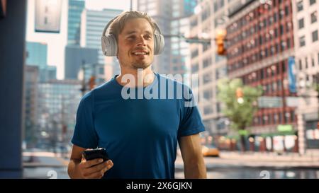 Handsome Young Man Walking on a Street of a Big City Center After Intense Fitness Workout and Using His Smartphone while Listening to Music on Hids Headphones. Urban Training Workout in the Morning. Stock Photo