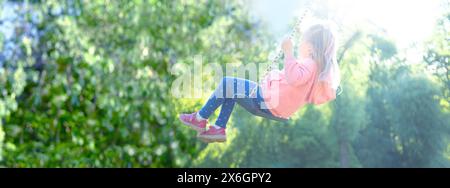 joyful little girl ascends into sunny sky on swing, blurred child figure in rays light, happy childhood, swing back and forth panorama Stock Photo