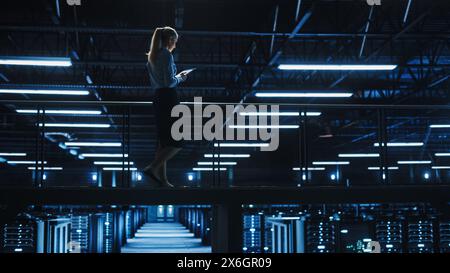 Female IT Specialist Using Tablet Computer in Data Center, Walking on a Bridge Overlooking Gigantic Server Farm Cloud Computing Facility. Succesful Businesswoman, e-Business Entrepreneur. Stock Photo
