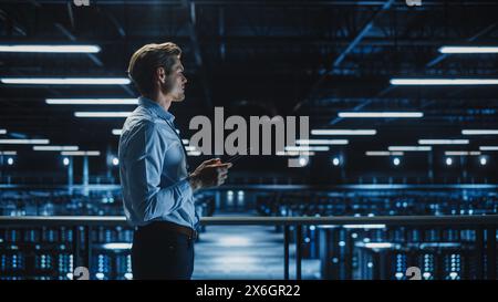 Handsome Smiling IT Specialist Using Tablet Computer in Data Center. Succesful Businessman and e-Business Entrepreneur Working in Big Server Farm Cloud Computing Facility. Stock Photo