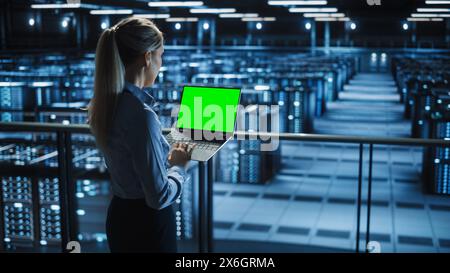 Female It Specialist Using Chroma Key Laptop in Data Center. Big Server Farm Cloud Computing Facility with Maintenance Engineer Working with Green Screen Computer. Stock Photo