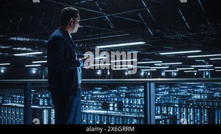 Handsome Smiling IT Specialist Using Tablet Computer in Data Center. Succesful Businessman and e-Business Entrepreneur Overlooking Server Farm Cloud Computing Facility. Stock Photo