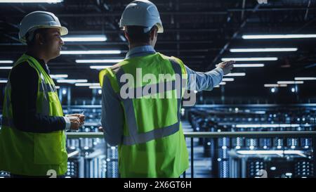 Data Center IT Specialist and System administrator Talk, Use Tablet Computer, Wearing Safety Wests. Server Clod Farm with Two Information Technology Engineers checking Cyber Security. Stock Photo