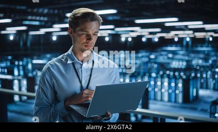 Handsome Smiling IT Specialist Using Laptop Computer in Data Center. Succesful Businessman and e-Business Entrepreneur Overlooking Server Farm Cloud Computing Facility. Stock Photo