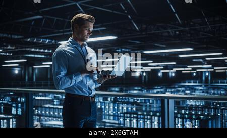 Handsome Smiling IT Specialist Using Laptop Computer in Data Center. Succesful Businessman and e-Business Entrepreneur Overlooking Server Farm Cloud Computing Facility. Stock Photo