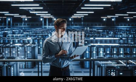 Portrait of IT Specialist Uses Laptop in Data Center. Server Farm Cloud Computing Facility with Male Maintenance Administrator Working on Cyber Security and Network Protection. Stock Photo
