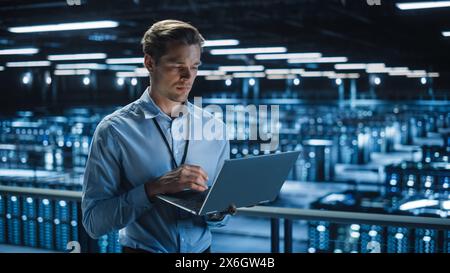 Handsome Smiling IT Specialist Using Laptop Computer in Data Center. Succesful Businessman and e-Business Entrepreneur Overlooking Server Farm Cloud Computing Facility. Stock Photo