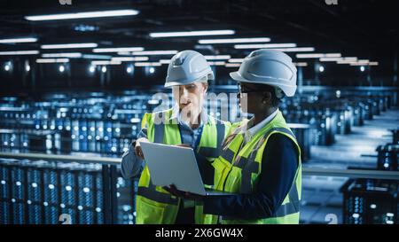 Data Center IT Specialist and System administrator Talk, Use Laptop Computer, Wearing Safety West. Server Cloud Farm Facility with Two Female Information Technology Engineers Doing Maintenance. Stock Photo