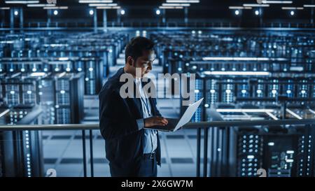 Portrait of IT Specialist Uses Laptop in Data Center. Server Farm Cloud Computing Facility with Male Maintenance Administrator Working. Cyber Security and Network Protection. Stock Photo