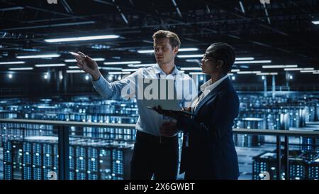 Data Center Female e-Business Enrepreneur and Male IT Specialist talk, Use Laptop. Information Technology Engineer and System Administrator work in Big Cloud Computing Server Farm. Stock Photo