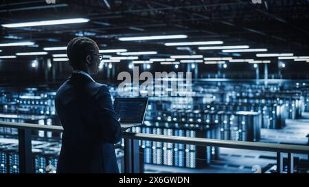 Female IT Specialist Using Laptop Computer in Data Center, Walking on a Bridge Overlooking Big Server Farm Cloud Computing Facility. Businesswoman, e-Business Entrepreneur. Stock Photo
