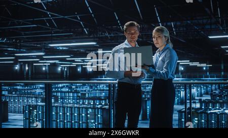 Data Center Female e-Business Enrepreneur and Male IT Specialist talk, Use Laptop. Two Information Technology Engineers work on Bridge Big Cloud Computing Server Farm. Stock Photo
