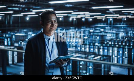 Handsome Smiling IT Specialist Using Tablet Computer in Data Center. Succesful Businessman and e-Business Entrepreneur Overlooking Server Farm Cloud Computing Facility. Stock Photo