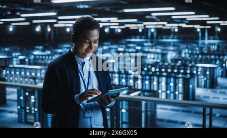 Handsome Smiling IT Specialist Using Tablet Computer in Data Center. Succesful Businessman and e-Business Entrepreneur Overlooking Server Farm Cloud Computing Facility. Stock Photo