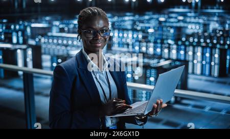 Data Center: Female Programmer Using Laptop Computer, Smiling and Looking at Camera. In Cloud Computing Server Farm System Administrator Working on IT Maintenance for Iaas, saas, paas. Stock Photo