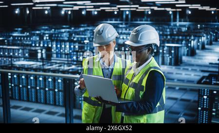 Data Center IT Specialist and System administrator Talk, Use Laptop Computer, Wearing Safety West. Server Cloud Farm Facility with Two Female Information Technology Engineers Doing Maintenance. Stock Photo