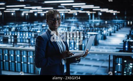 Data Center: Female Programmer Using Laptop Computer, Smiling and Looking at Camera. In Cloud Computing Server Farm System Administrator Working on IT Maintenance for Iaas, saas, paas. Stock Photo