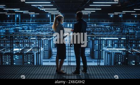 Data Center Male It Specialist and Female e-Commerce Manager Talk, Use Laptop. Big Server Farm Cloud Computing Facility with Information Technology Professionals Working. Stock Photo
