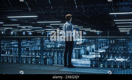 Handsome Smiling IT Specialist Using Tablet Computer in Data Center. Succesful Businessman and e-Business Entrepreneur Overlooking Big Server Farm for Cloud Computing. Stock Photo