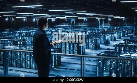 Handsome Smiling IT Specialist Using Tablet Computer in Data Center. Succesful Businessman and e-Business Entrepreneur Overlooking Server Farm Cloud Computing Facility. Stock Photo