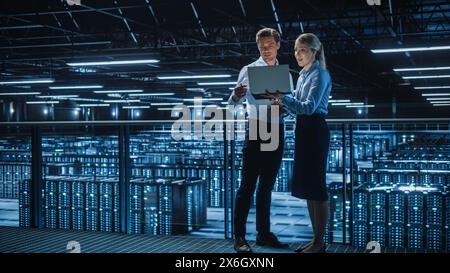 Data Center Female e-Business Enrepreneur and Male IT Specialist talk, Use Laptop. Two Information Technology Engineers work on Bridge Big Cloud Computing Server Farm. Stock Photo