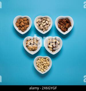 Top view, close up, on a plain blue background, six white heart shaped bowls with shelled mixed dried fruit. Stock Photo